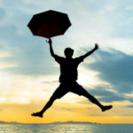 Man in silhouette celebrating by jumping holding an umbrella over water at dusk